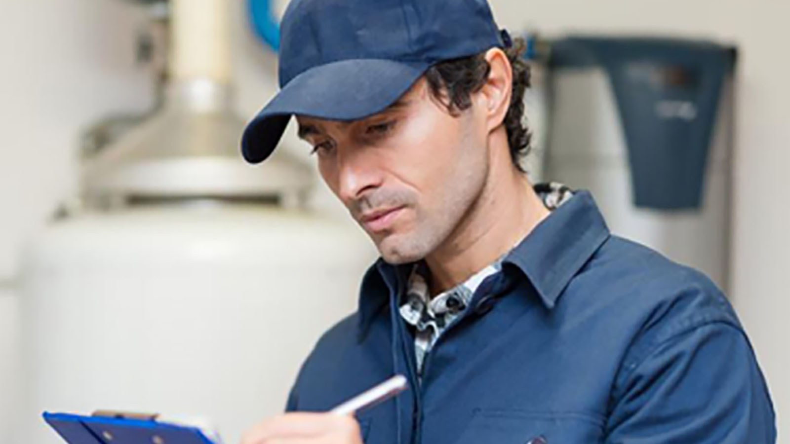 Building inspector in blue coveralls and hat taking notes on clipboard.