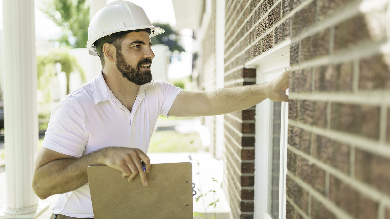 A man with a white hard hat holding a clipboard, inspecting a brick house.
