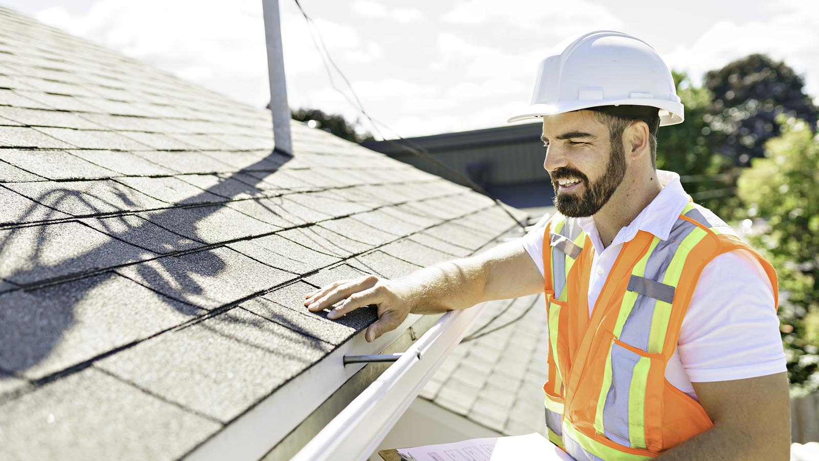 A building inspector in a white hard hat and orange high-vis vest, inspecting a tiled roof.