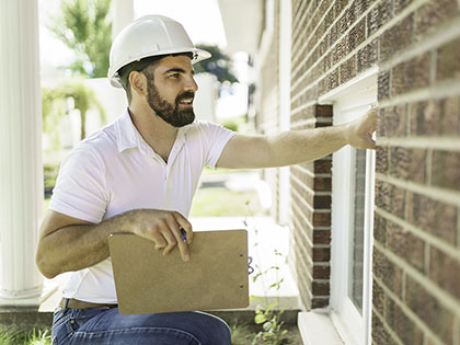 A man with a white hard hat holding a clipboard, inspecting a brick house.