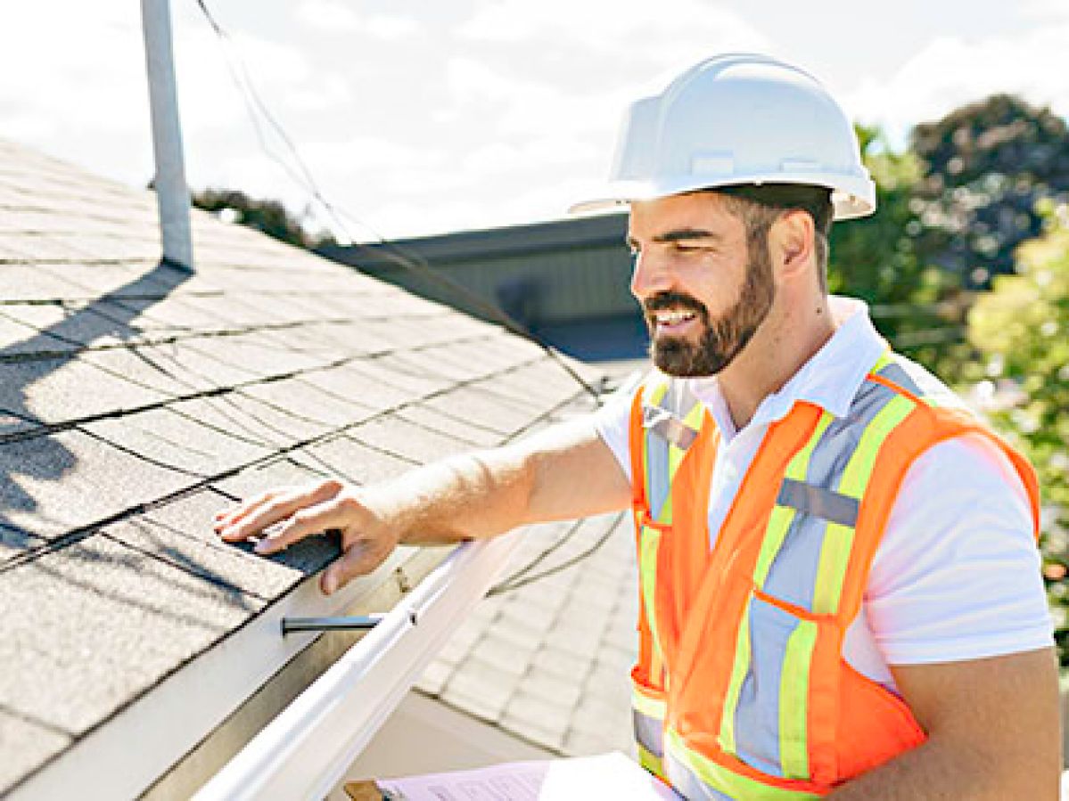 A building inspector in a white hard hat and orange high-vis vest, inspecting a tiled roof.