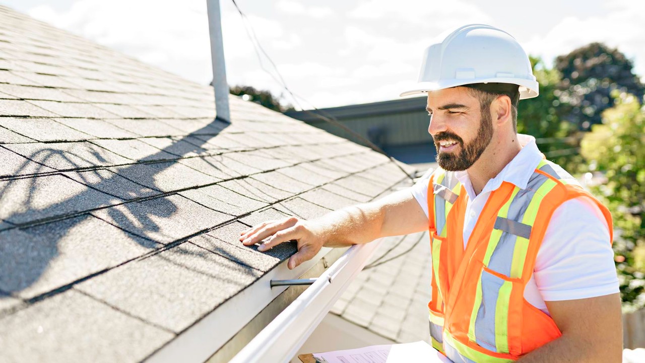 A building inspector in a white hard hat and orange high-vis vest, inspecting a tiled roof.