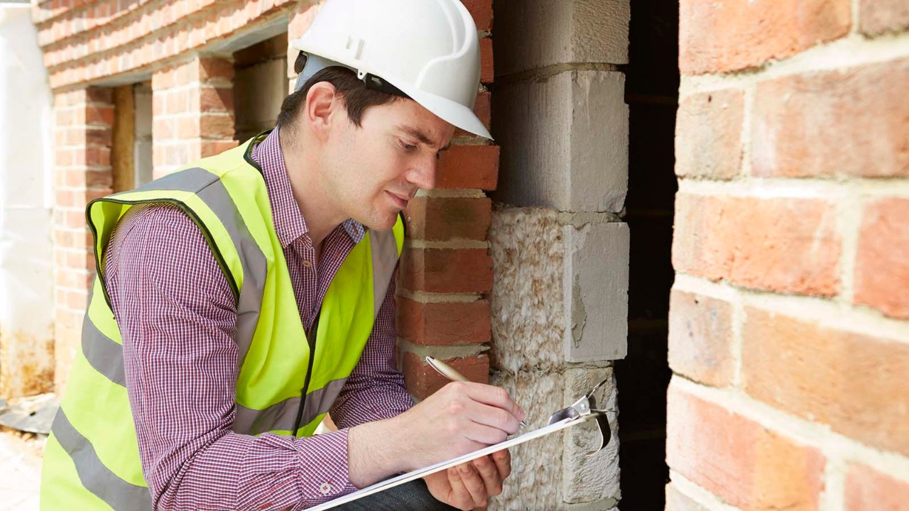 Building inspector in high-vis vest and hard hat taking notes on clipboard.